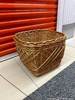 Wicker cloth basket with visible woven pattern, thicker braided rim, placed on tiled floor with orange storage door background.