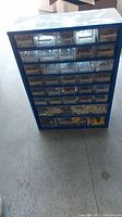 Full front view of blue metal hardware cabinet with 33 clear drawers showing some contents inside including screws and miscellaneous hardware
