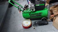 Full view of green Kawasaki air compressor with attached handle, wheels, black coiled air hose, and red and yellow hose reel on floor beside compressor.
