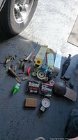 Photo showing assorted fishing spoons, fishing knife with sheath, fishing line spools, fishing weights, two bobbers, handheld scale, and assorted fishing hooks and tools laid out on floor near tire.