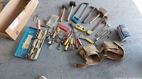 Overall view of mixed tools laid on floor showing screwdrivers, hammers, wrench, socket set, and leather tool belt.