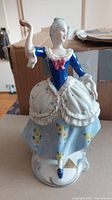 Front view of Royal Dux bone china figurine depicting a dancing lady in 18th century style dress, blue and white with floral designs, standing on a decorated base.