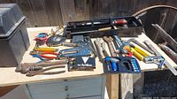 Assorted hand tools laid out on wooden table outside including pliers, clamps, wrenches, screwdrivers, measuring tape, lock pick set, wire stripper.