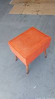 Full view of vintage orange upholstered sewing stool with decorative stitching on seat, standing on concrete floor.