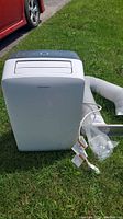 Full view of white Garrison portable air conditioner on grass with exhaust hose and accessories laid out beside it.