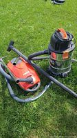 Ridgid shop vac with orange top and flexible hose, and Kenmore red vacuum cleaner with HEPA filter, both on grass outdoors.
