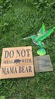 Wooden 'Do Not Mess With Mama Bear' sign and green hummingbird decoration on grass