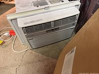 Full view of white Frigidaire window mount air conditioner unit sitting on the floor with power cord visible.
