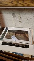 Stack of multiple unframed art prints placed on a table against a concrete wall, showing varied artwork including a maritime scene and portrait.