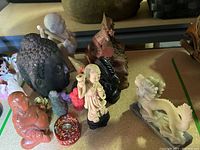Photo showing multiple Asian figurines including Laughing Buddha, Buddha head, dragon, miniature Buddhas, and a girl figurine displayed on a table.