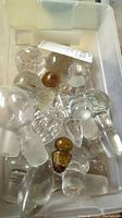 Container filled with multiple clear glass bottle stoppers of different shapes and sizes, including a few amber glass ones.