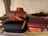 Vintage Argus 35mm camera in brown leather case on black case, with small wooden and burgundy boxes nearby, on a surface.