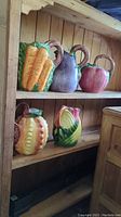 Five vegetable-shaped ceramic pitchers placed on wooden shelves; includes carrot, eggplant, tomato, corn, and cabbage designs.
