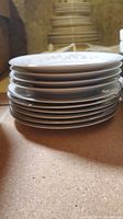 Stack of white ceramic dinner plates with light grey floral trim around scalloped edges, stacked vertically, some chips and crazing visible.