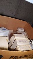 Cardboard box containing multiple boxed porcelain dinnerware items, some unwrapped pieces visible, cardboard labeled FRAGILE