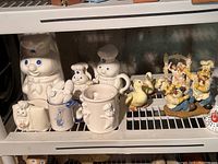 Shelf with assorted Pillsbury Doughboy ceramic items including large cookie jar, mugs, napkin holder, and a smaller figurine. Also includes duck and chicken ceramic figurines and colorful baker men figures.