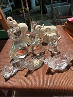 Photo showing a collection of glass animal figurines including cat, dog, hippo, and two white ceramic elephants on a woven surface.