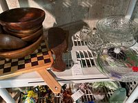 Wooden bowls stacked with a wooden pizza paddle and checkered wooden cheese board, including several glass serving bowls and plates on a white shelf.
