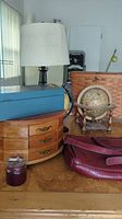 Photo showing lamp with beige shade, wooden jewelry box with three drawers, blue metal lock box, decorative globe with wooden stand, basket and dark red pocketbook on table.