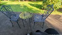 Full view of cast iron patio furniture set including round glass top table and two matching chairs on concrete pad.