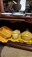 Yellow pottery and bakeware including polka dot hen-shaped covered dish, baking dishes, canisters, and platter shown on wooden surface in basement setting.