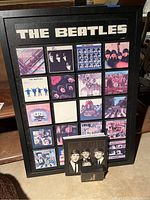 Framed Beatles collage with 20 album cover images, two 8-track cartridges, and a small book placed in front, showing wear and peeling labels on cartridges.