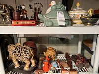Overview of items on two shelves in basement: Italian tureen, carved elephant, green turtle ceramic figure, vintage man and dog carvings, brass dishes, wooden figures, ceramic jars.