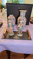 Photo showing two clown figurines and two vases on a table, with a black framed object behind as background.
