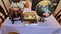 Vintage Replogle Starlight globe seated on metal base, two dark wood Asian-style vase stands, Asian wooden framed wall art, fringed leather ladies shoulder bag on table with light purple cloth.