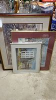 Four framed cityscape prints leaning against each other on a basement floor with glassware on table behind