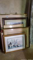 Stack of framed artworks with visible front frame showing four fishing boats in harbor.