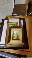 Stack of six diverse framed artworks and frames mainly of landscape paintings, showing different frame styles and some wear.