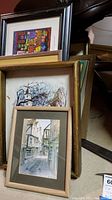 Group of multiple framed wall art prints leaning against a piece of furniture. Scenes feature cityscapes and houses in different styles. Frames vary in color and style.