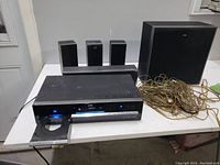 Black JVC home stereo system with four satellite speakers, a large subwoofer, receiver unit on white table. Wires bundled on right.