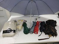 Seven umbrellas in different colors and patterns laid out on a white table, including a large gray umbrella, green, blue patterned, red, black, brown with strap, and beige umbrella.