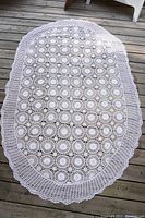 White oval crocheted and macrame tablecloth spread out on wood deck showing circular motifs and scalloped edges.