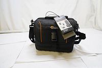 Front view of black Lowepro Adventura SH160 II camera bag with orange accents and attached product tag, showing main flap with buckle and shoulder strap.