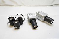 Two Samsung surveillance cameras with lenses attached and three interchangeable lenses. Cameras have silver bodies with Samsung logos and black lenses attached, additional lenses displayed separately.
