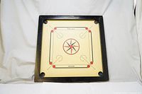Front angle view of the square wooden carrom board showing black frame and cream playing surface with red and black design, corner pockets visible.