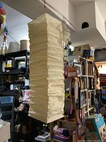 Side view of rectangular cream-colored paper lamp shade showing wavy drape texture, hanging indoors in a room with cluttered shelves.