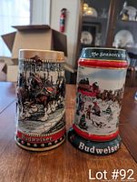 Photo of two Budweiser collectible beer steins, one from 1988 featuring a festive holiday harness scene, and a second from 1991 with a winter scene with Clydesdales and wagon.