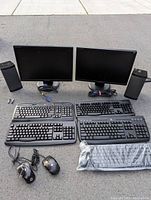 2 black Samsung flat screen monitors with VGA cables, 4 black wired keyboards, 2 black wired computer mice, and 2 tall black desktop speakers on a gray surface.