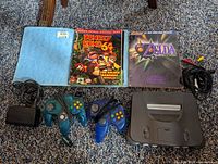 Photo shows Nintendo 64 console, two controllers (one original translucent teal, one aftermarket translucent blue), power supply, composite AV cable, and two player strategy guides for Donkey Kong 64 and Zelda: Majora's Mask laid out on carpet.