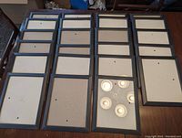 All 23 black 8x10 picture frames arranged in rows on a wooden table, some showing backing boards with hanging hardware, one with a reflective glass pane.