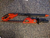 Black & Decker cordless pole hedge trimmer laid flat, showing the handle, telescoping pole, and blade assembly with battery compartment.