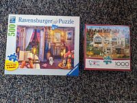 Photo of two boxed jigsaw puzzles on carpet: Ravensburger 500-piece with cozy indoor scene and dog, and Buffalo 1000-piece Charles Wysocki house puzzle.