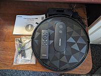 Photo showing Airrobo P20 robot vacuum with geometric patterned top, remote control, charging dock with power adapter, extra parts in sealed bag, and user manual placed on wooden surface.