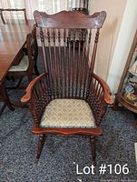 Front-facing full view of an antique wooden rocking chair showing carved scroll design on the top backrest, spindle back, wooden armrests, and patterned upholstered seat cushion. Chair is placed on a carpeted floor near a dining table and wall.