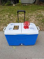 Full frontal image of a large Rubbermaid cooler outside on grass. Cooler is blue with white lid that splits in half with four built-in cup holders, black extendable handle pulled up, and side carrying handles visible.