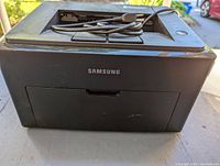Front view of the black Samsung laser printer showing the Samsung logo and the front paper output area with some minor surface scratches.
